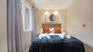 A bedroom with a bed, grey curtains, a window, a wall-mounted artwork, and a bedside table with a lamp.