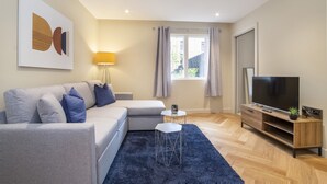 A modern living room with a grey sofa, a small white side table, a blue rug, a wooden TV stand, and a large window with curtains.