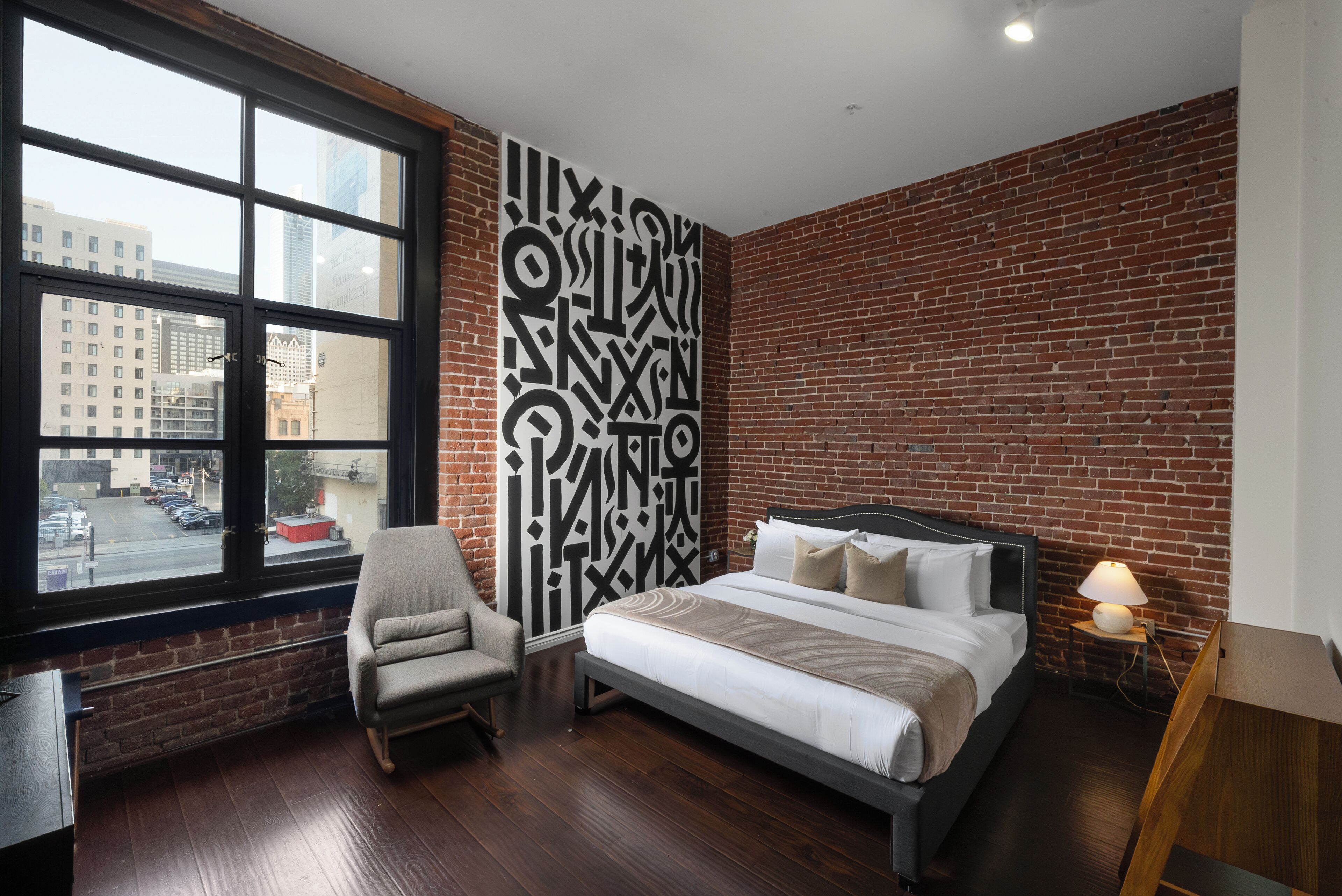 Una habitación de hotel moderna con pared de ladrillo, una ventana grande, una cama, una silla y una mesita de noche con lámpara.