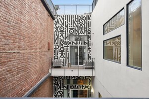 Uma viela estreita com uma parede de tijolos de um lado e um edifício branco com padrões geométricos do outro. Há uma pequena varanda com grades de metal e algumas cadeiras.