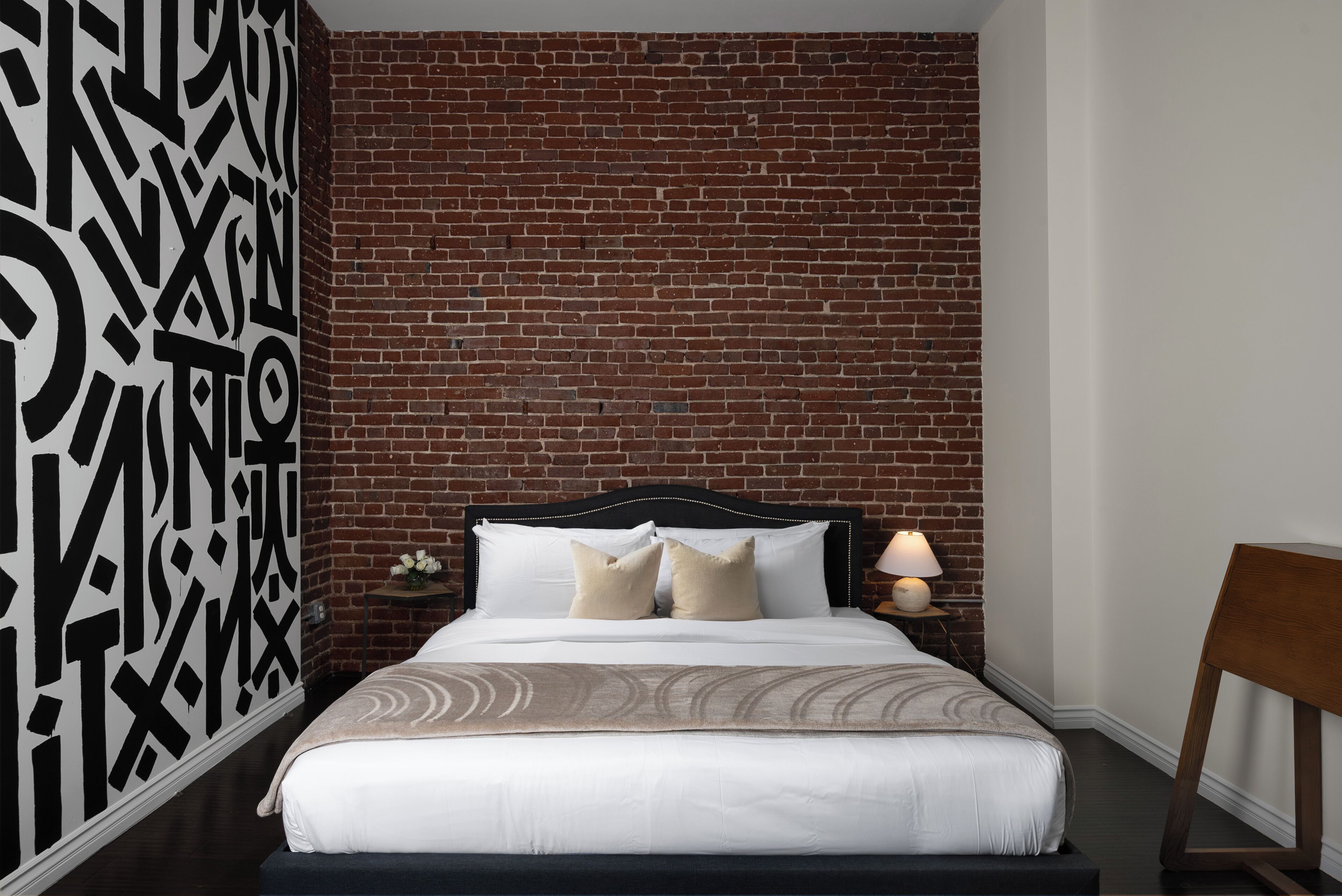 A bedroom with a brick wall, a bed with white bedding, two pillows, a nightstand with a lamp, and a wooden chair.