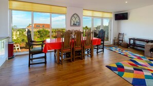 Un comedor con una mesa y sillas de madera, ventanas amplias con persianas, una alfombra colorida y un televisor en la pared.