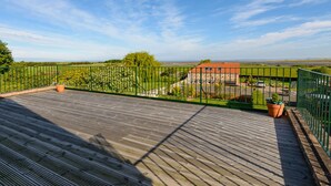 Terraza de madera con barandilla de metal verde, con vistas a una zona cubierta de césped, un edificio de piedra y un estacionamiento.
