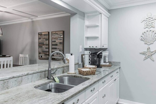 A modern kitchen with a granite countertop, stainless steel sink, and white cabinetry. There are decorative wall art pieces and a coffee maker on the counter.