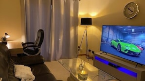 Ein modernes Wohnzimmer mit einem Ledersofa, einem Glastisch, einem Fernseher, der einen Sportwagen zeigt, und einer Lampe.