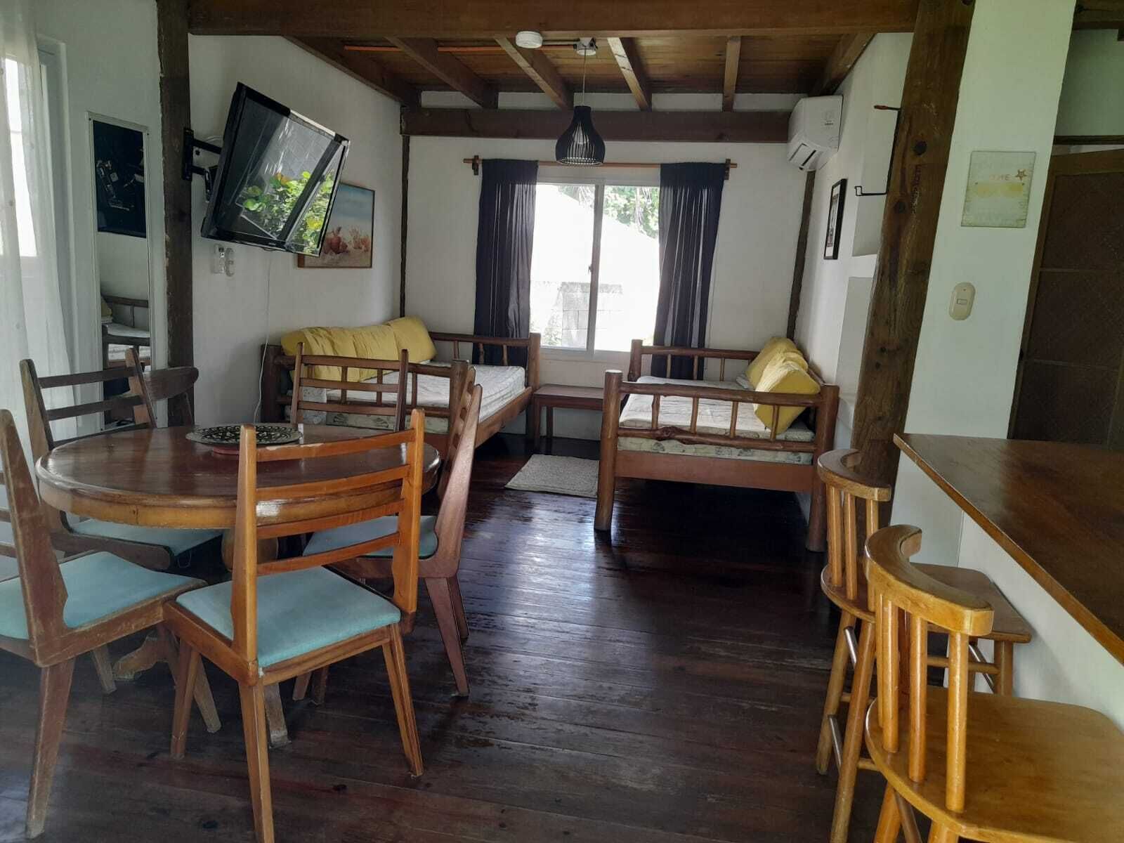 Ein rustikaler Essbereich mit einem runden Tisch und Stühlen, zwei Holzbetten mit Kissen, einem an der Wand montierten Fernseher und einem Fenster mit Vorhängen.