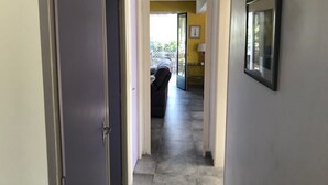 Un couloir avec une porte à gauche, un tableau encadré à droite, et, au fond, on aperçoit une pièce avec un lit et une fenêtre.