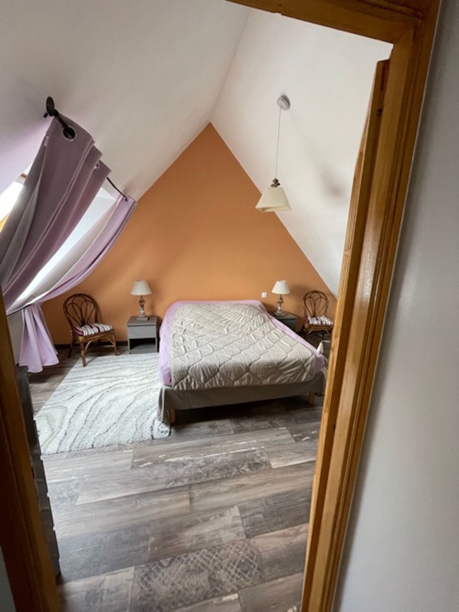 Une chambre avec un plafond en pente, un lit, deux tables de chevet avec des lampes, une chaise et une grande fenêtre avec des rideaux.