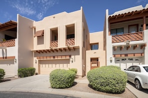 A modern two-story building with a beige facade, a wooden balcony, and a white car parked in the driveway.