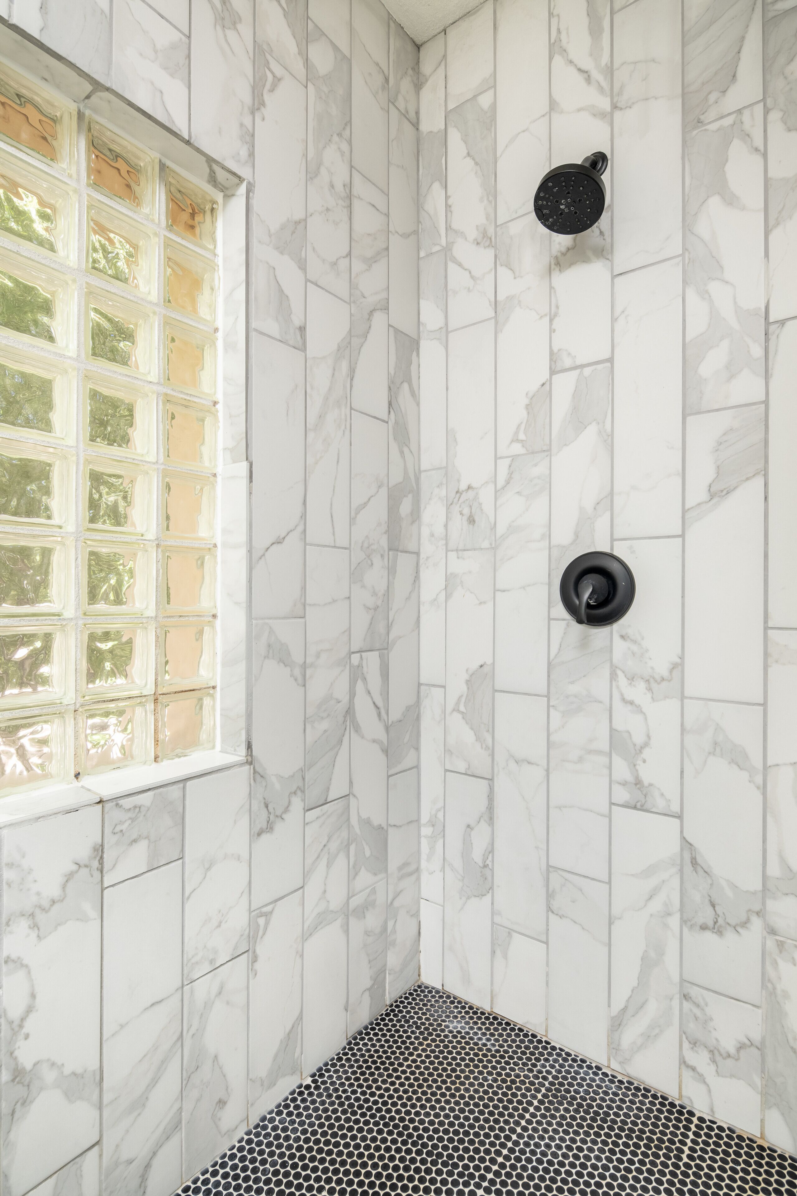 A bathroom with marble walls, a shower with a black head, and a window with a grid pattern.