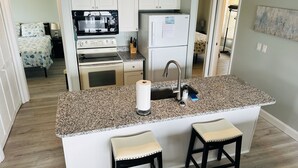 Eine kompakte Küche mit Granitarbeitsplatte, integriertem Backofen, Mikrowelle und Kühlschrank. Zwei Barhocker und ein Spülbecken mit Wasserhahn sind vorhanden.