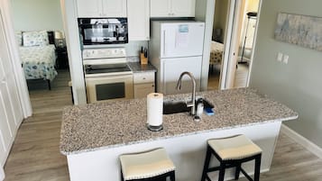 A compact kitchen with a granite countertop, a built-in oven, a microwave, and a refrigerator. There are two bar stools and a sink with a faucet.