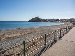 A coastal promenade with a pebbled beach, a protective railing, and a distant hill with buildings. - Ty Jem (Criccieth)
