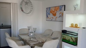 A modern dining area with a marble table, white upholstered chairs, a chandelier, and a fish tank on a white shelf. - Luxurious Apartment 4to level with security camera (Santo Domingo Norte)