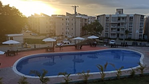 A swimming pool with surrounding lounge chairs and umbrellas, surrounded by a fence, with buildings in the background. - Luxurious Apartment 4to level with security camera (Santo Domingo Norte)