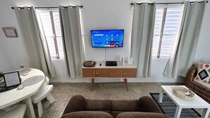 A modern living room with a TV, a sofa, a dining table, and a small white table with a decorative item.