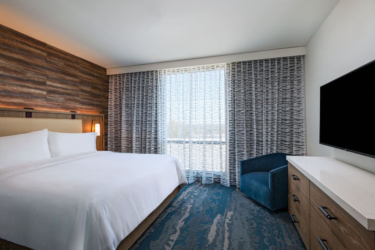 Een hotelkamer met een groot bed, een flatscreen televisie, een houten hoofdbord, een blauwe fauteuil en een gordijn met dessin.
