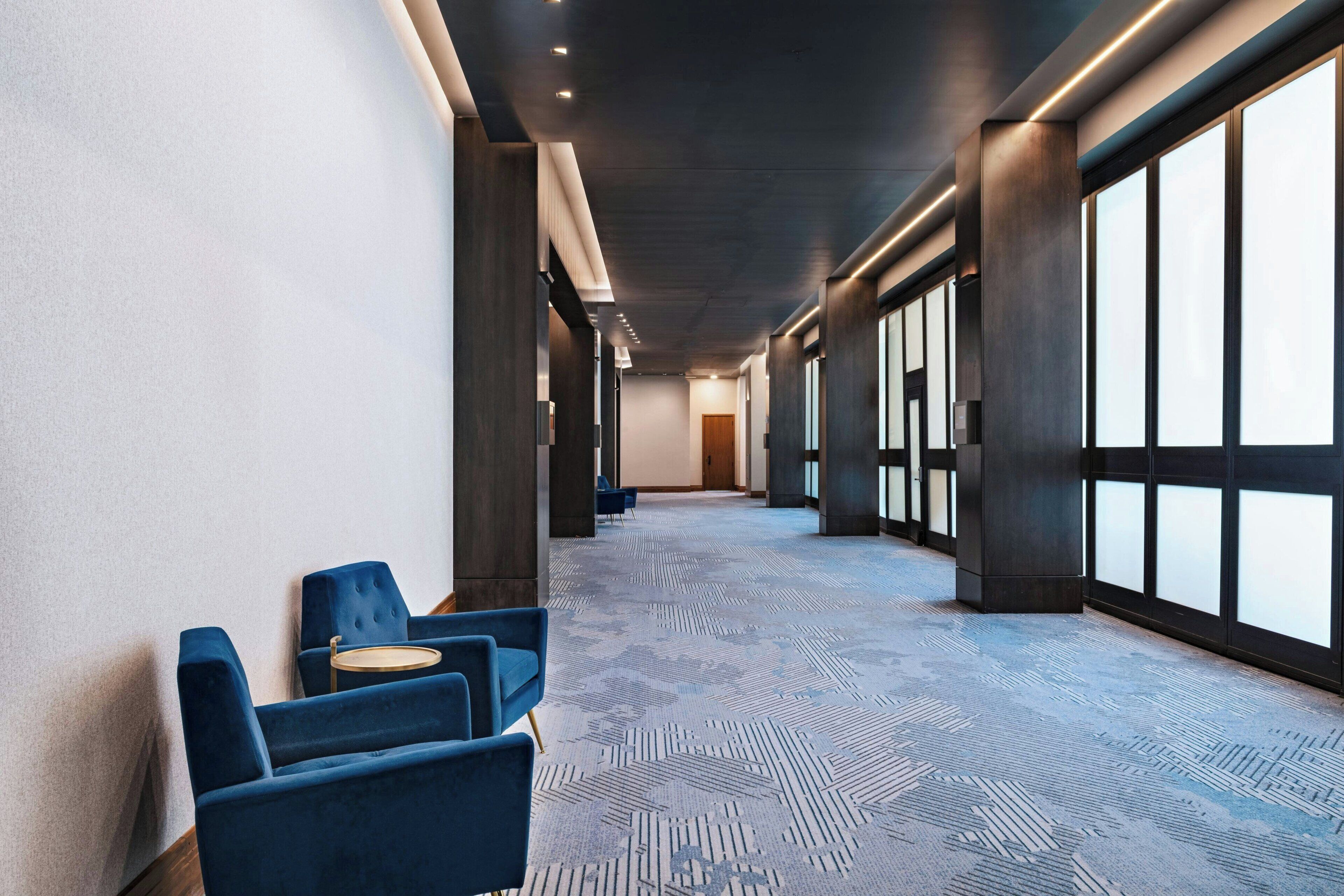 A hotel corridor with a patterned carpet, blue upholstered chairs, and large windows.