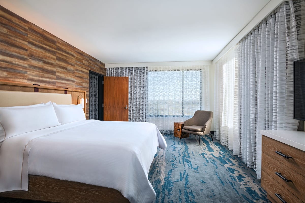 Hotelkamer met een groot bed, een houten hoofdbord, een klein nachtkastje, een stoel, een televisie en een raam met dunne gordijnen.