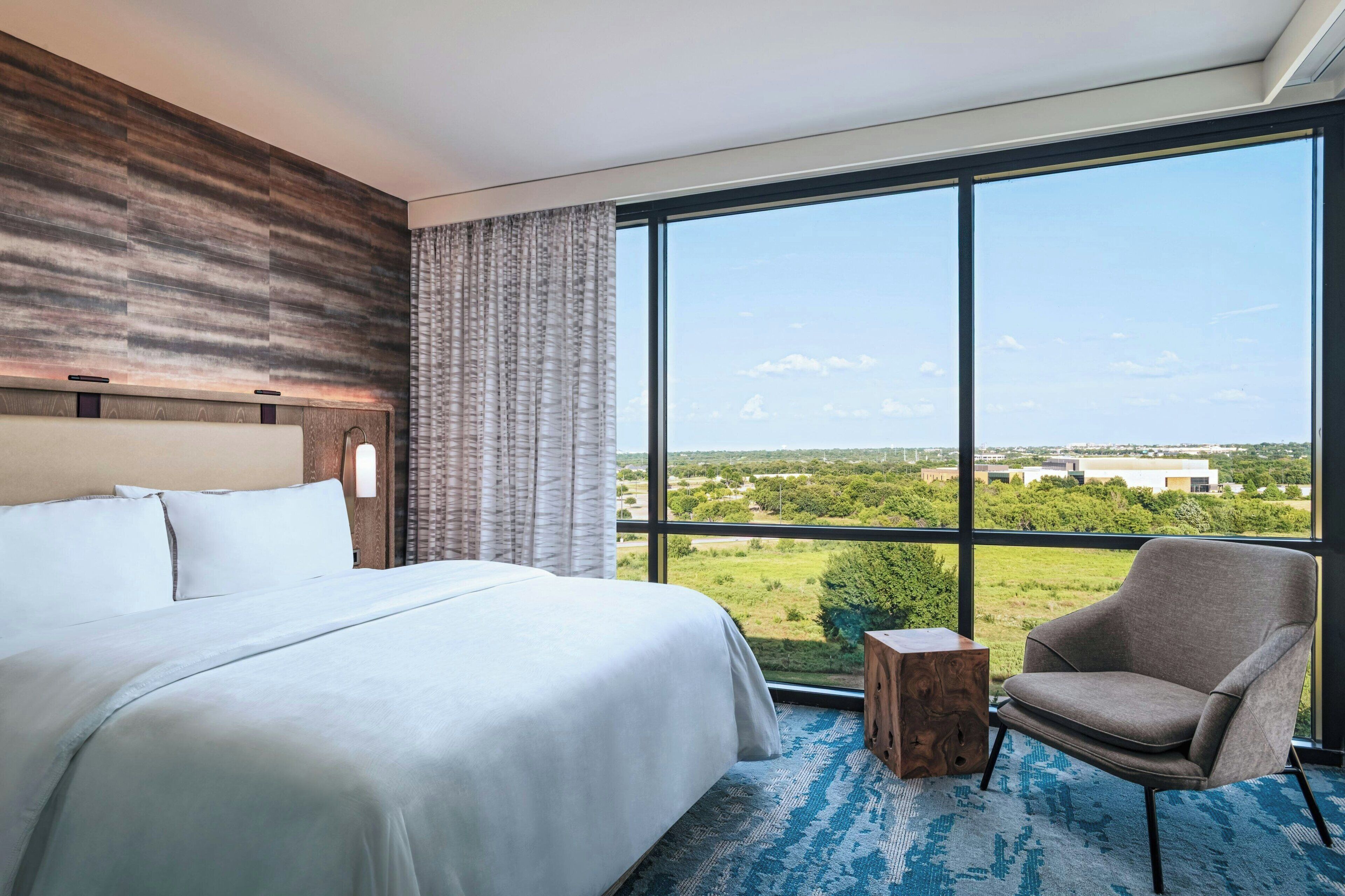 A hotel room with a large bed, a chair, a small wooden side table, and a view of the countryside through large windows.