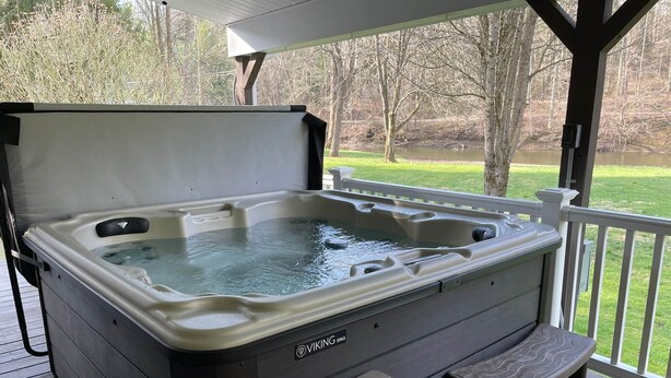 A hot tub with a wooden deck, a covered area, and a view of a grassy field and trees. - Riverside Escape - Riverfront Home on 1 acre (Hanover Township)