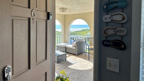 Una puerta que da a un balcón con vistas a la playa, un cartel de 'No Fumar' y letreros decorativos que dicen 'The Beach is My Happy Place'.
