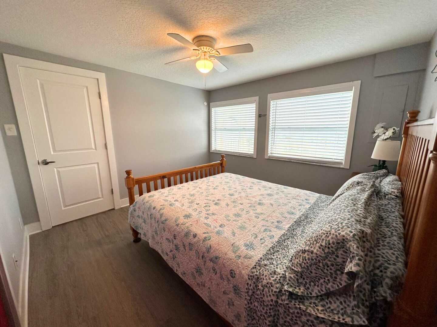 Un dormitorio con cama, ventilador de techo, dos ventanas con persianas y una puerta.