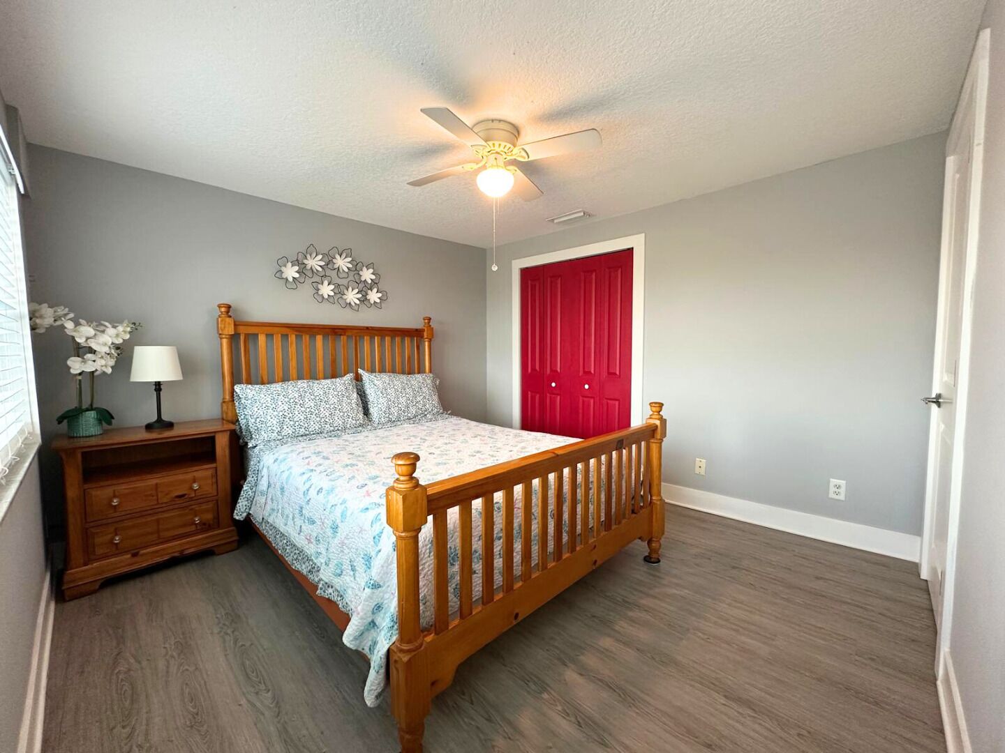 A bedroom with a wooden bed, a nightstand, a lamp, a ceiling fan, a red door, and a wall decoration.