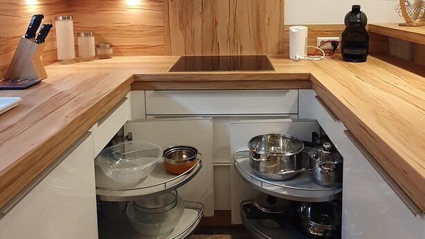 Una cocina con encimeras de madera, armarios blancos y horno empotrado. Se observan dos estantes con vajilla y un pequeño florero con plantas secas.