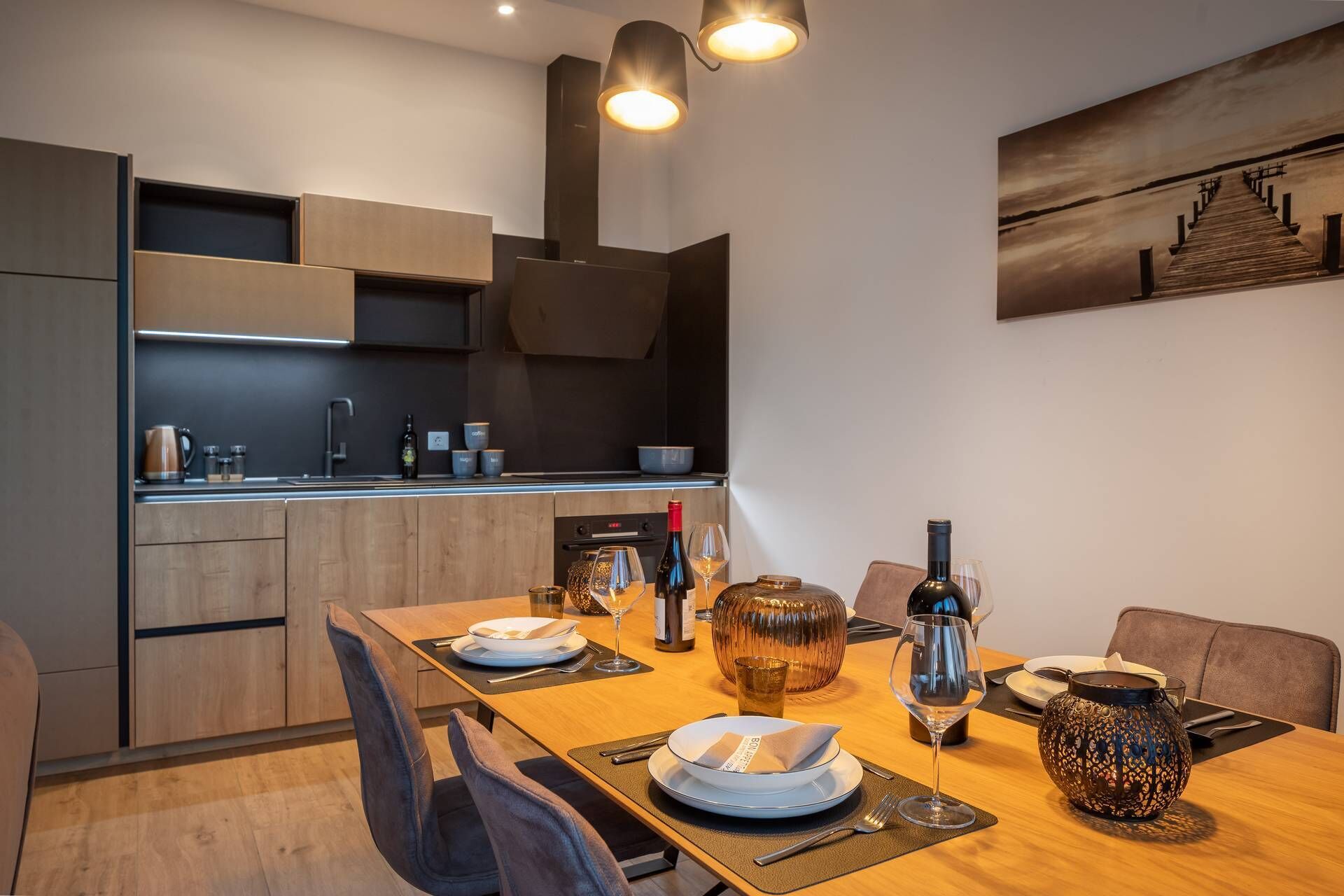 A modern kitchen with wooden cabinets, a dining table set for two, and a painting on the wall.
