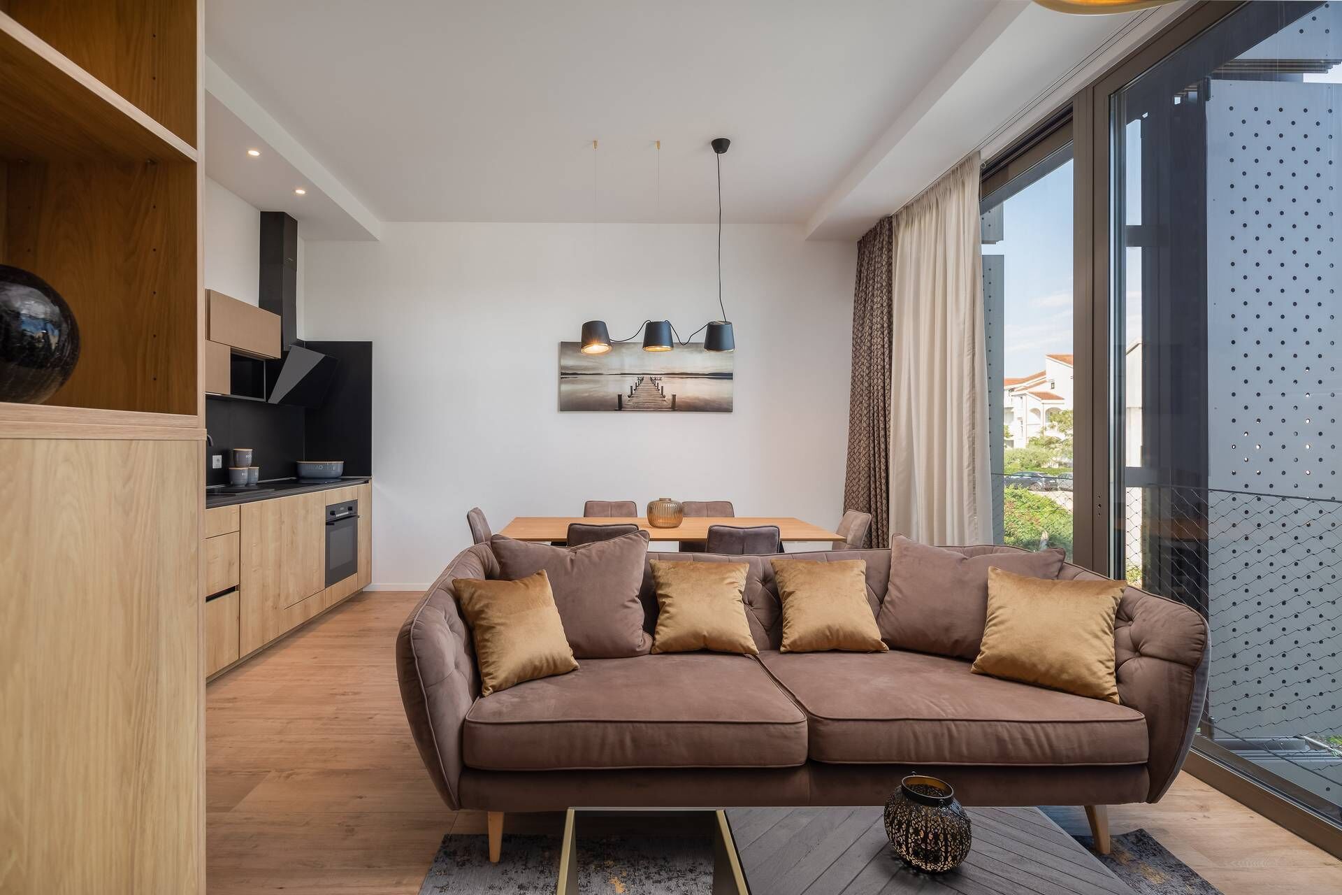 A modern living room with a brown sectional sofa, a dining table, and a kitchen area with wooden cabinets and a black stove.