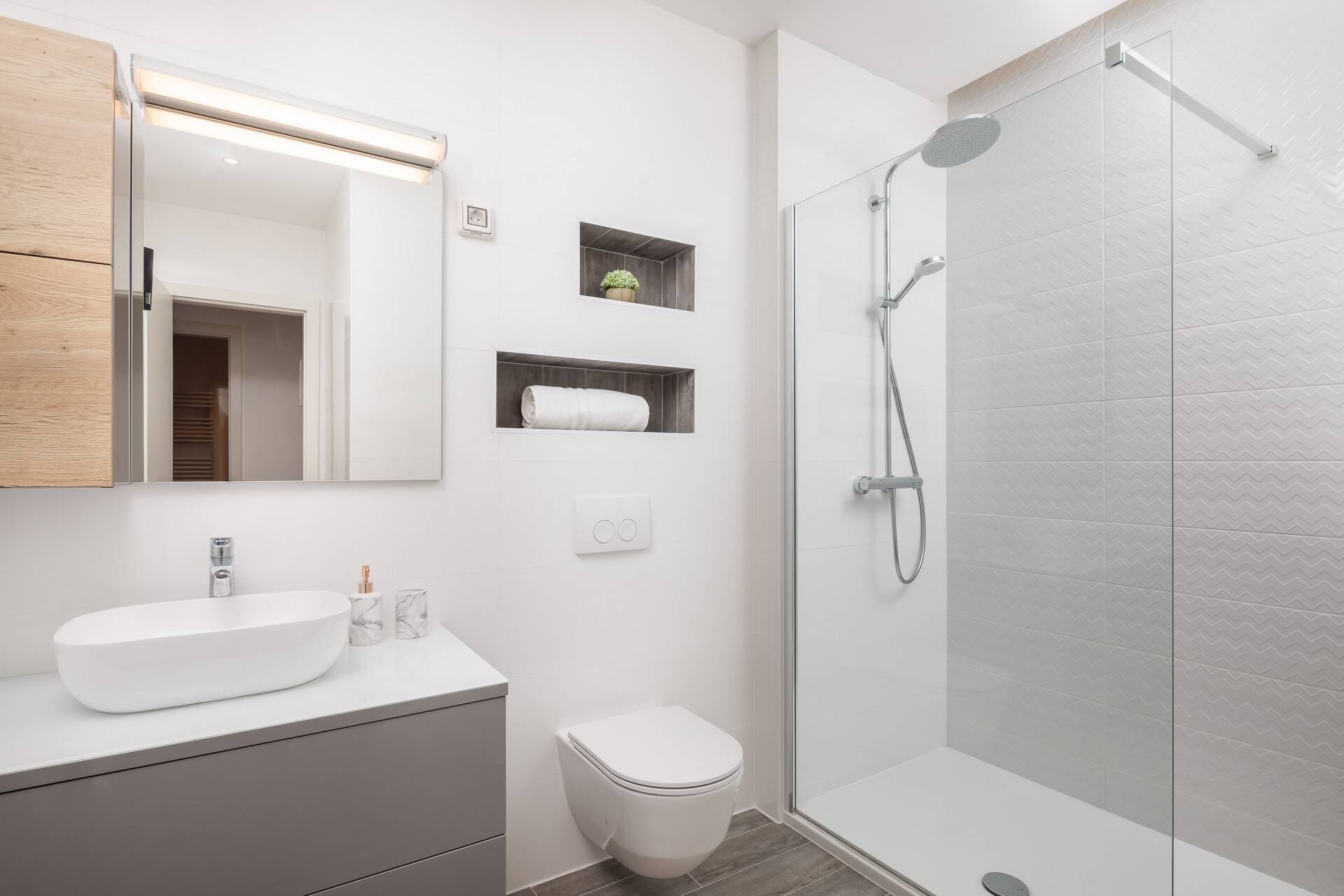 A modern bathroom with a white sink, toilet, and shower. There is a mirror, a small shelf with a plant, and a light fixture above the mirror.