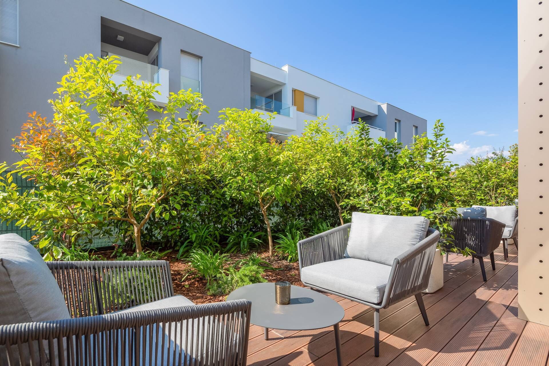 An outdoor patio with wicker furniture and a small round table, surrounded by lush greenery and a modern building in the background.