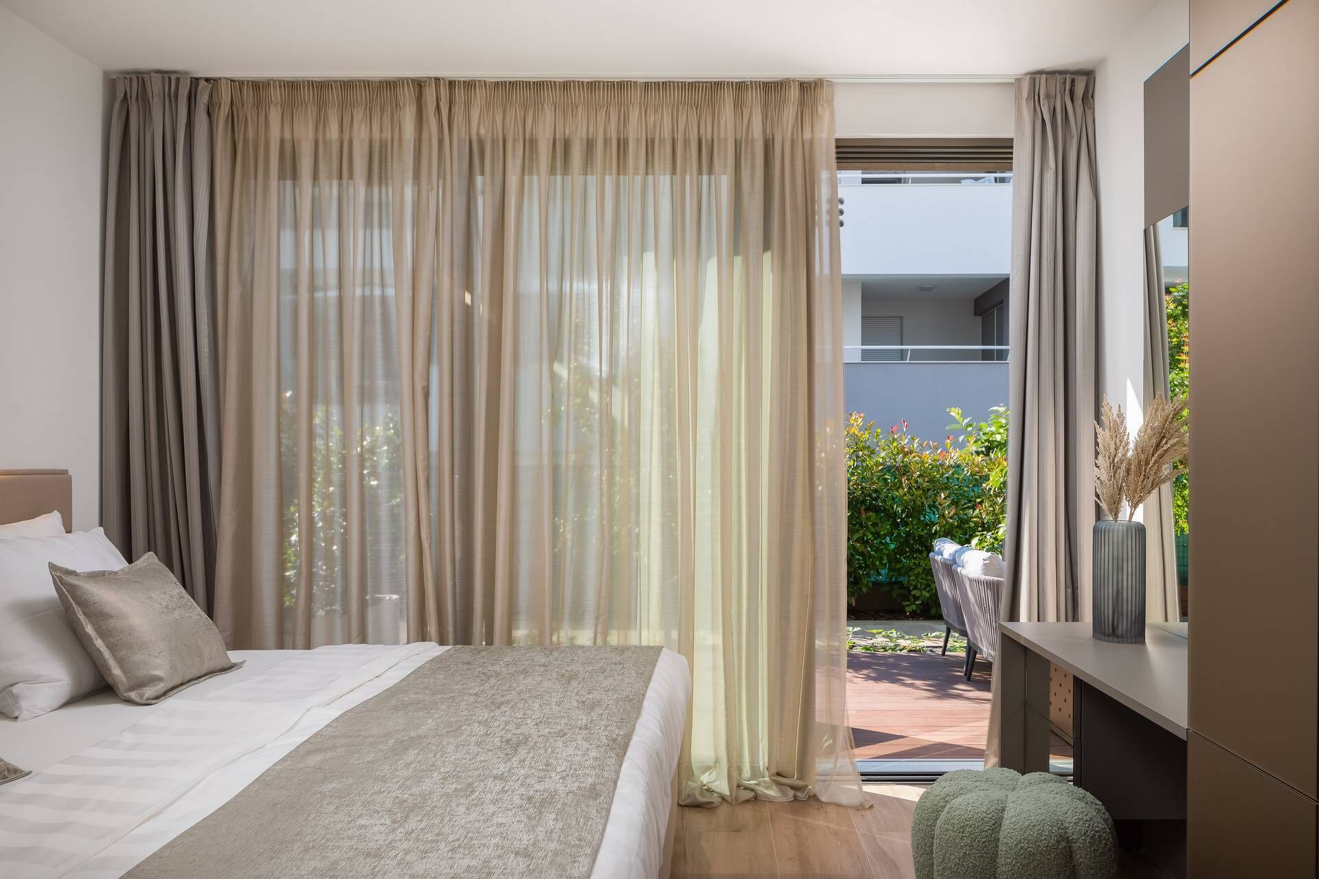 A modern bedroom with a large bed, a desk, and a view of greenery outside.