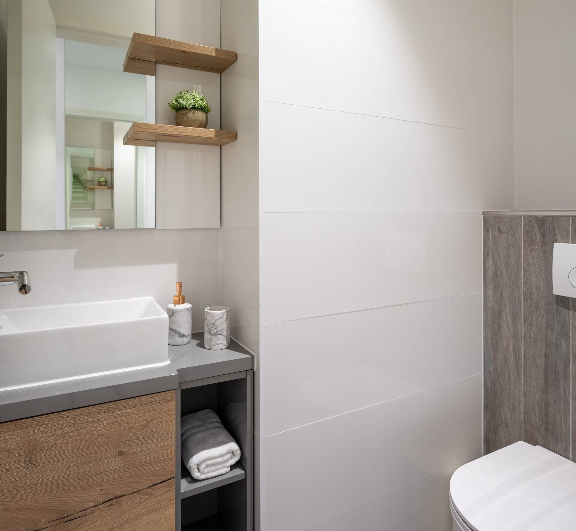 A modern bathroom with a white sink, marble containers, a towel rack, and a wooden shelf with a plant.