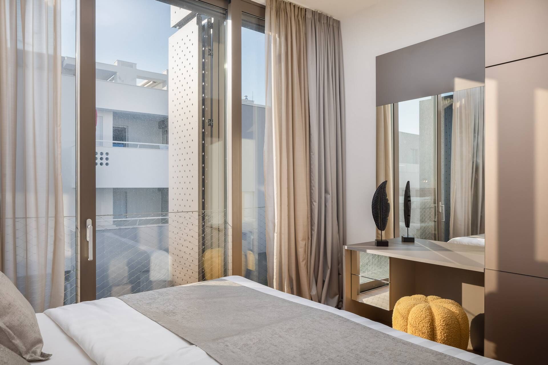 Une chambre d’hôtel moderne avec un grand lit, une petite table, un miroir et une vue sur la ville grâce à de grandes fenêtres du sol au plafond.