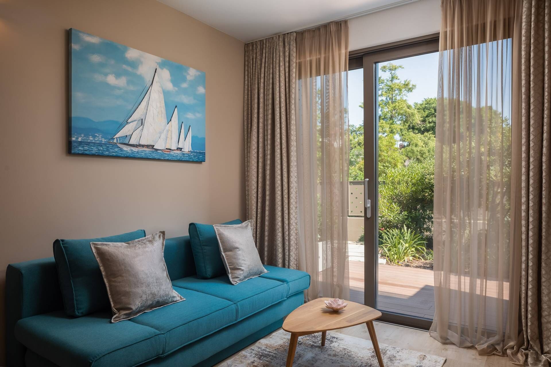 A living room with a blue sofa, a painting of sailboats, and a view through large glass doors.