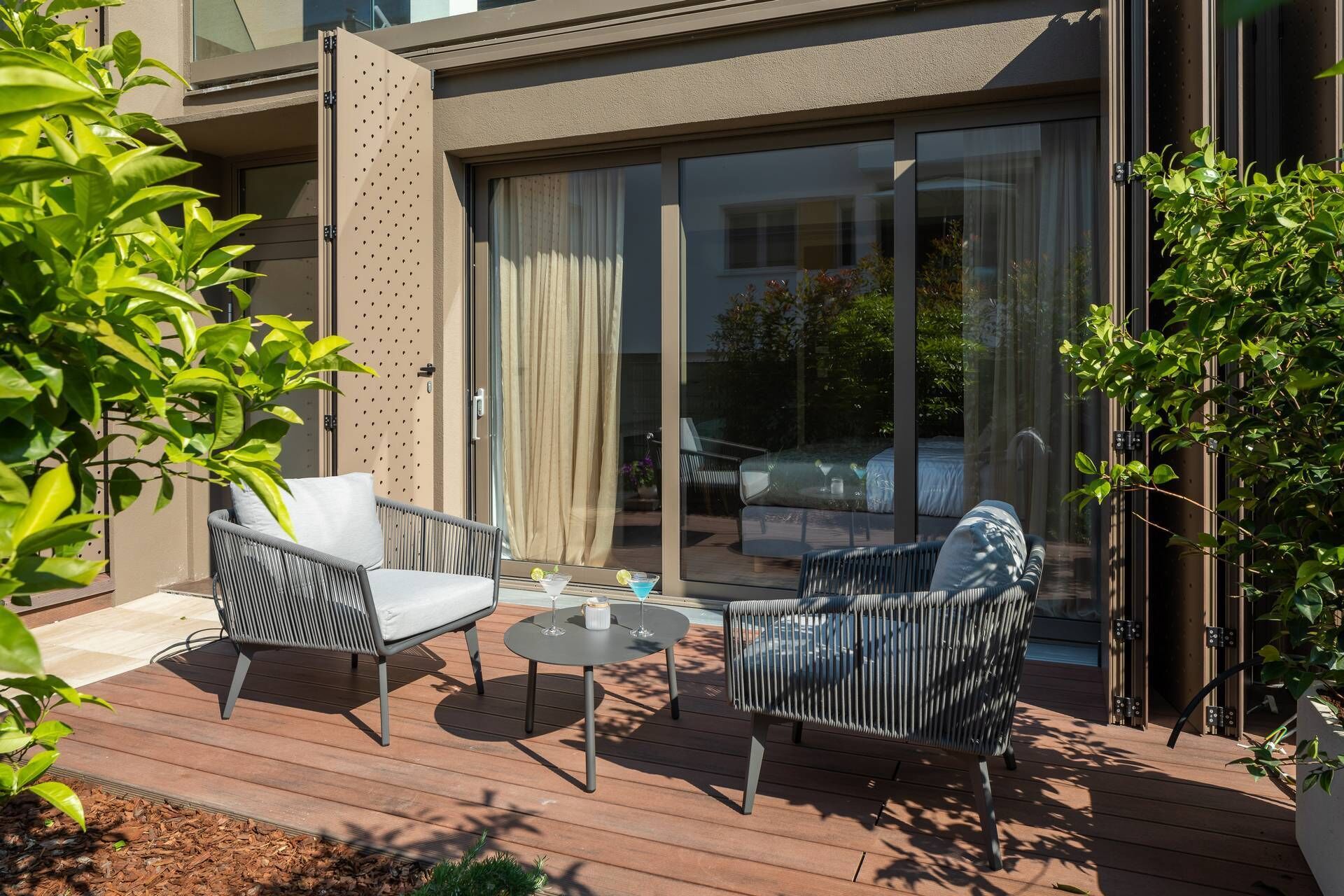 A patio with two wicker chairs and a small round table, a sliding glass door, and a view of a garden.