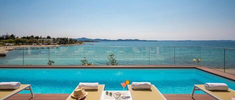 Ein Poolbereich mit Liegestühlen, einem Tisch mit Getränk und Blick auf das Meer und ferne Berge.
