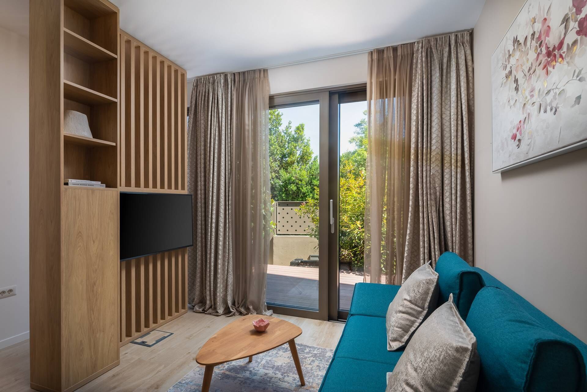Ein modernes Wohnzimmer mit einem türkisfarbenen Sofa, einem Holzregal, einem Flachbildfernseher und einer Schiebeglastür, die zu einem Außenbereich führt.