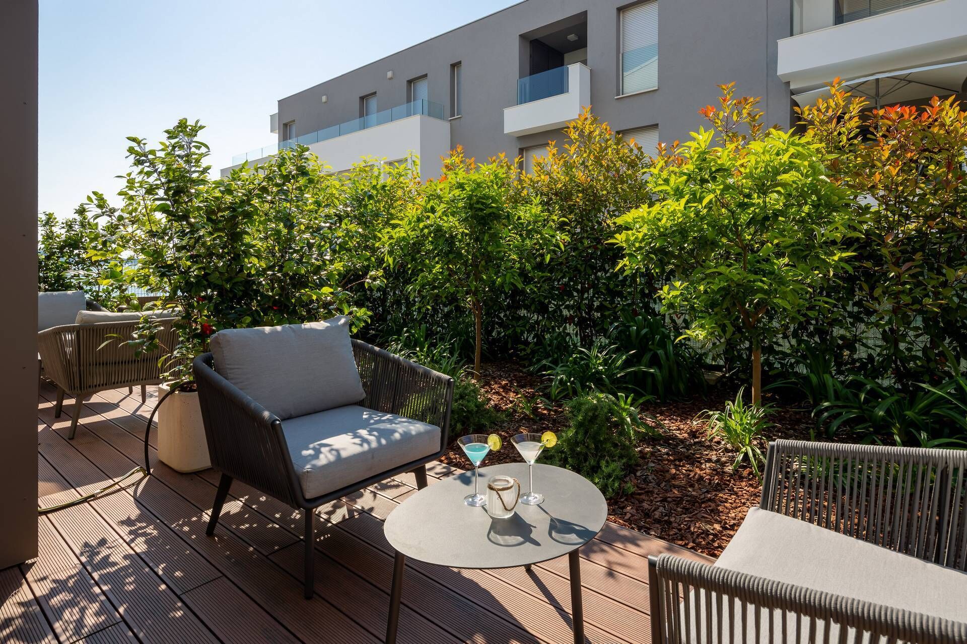 Un'area relax esterna moderna con mobili in vimini e un tavolo rotondo, circondata da vegetazione lussureggiante e un edificio sullo sfondo.