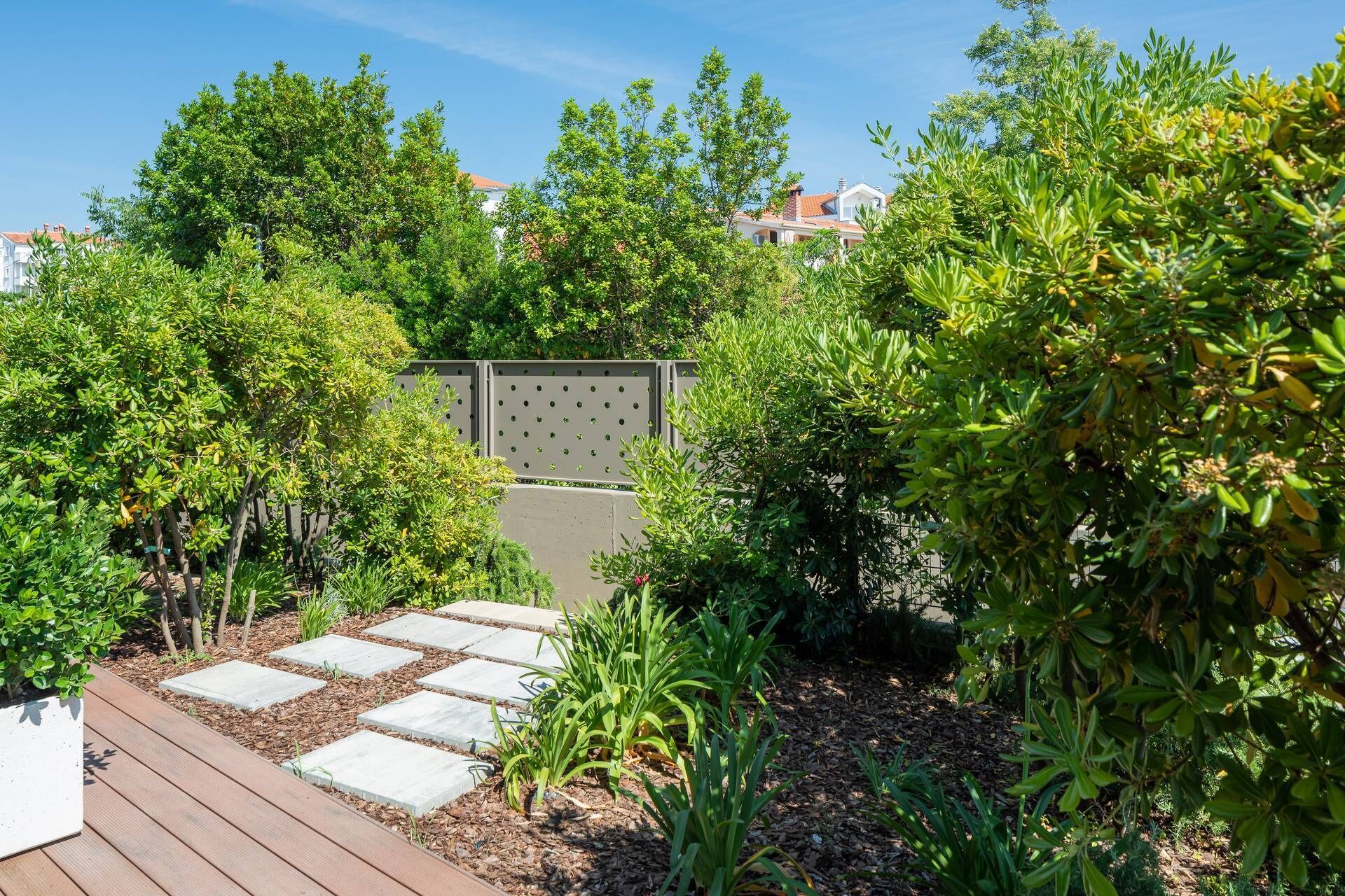 Een tuin met een houten terras, een pad van stenen en diverse groene planten.