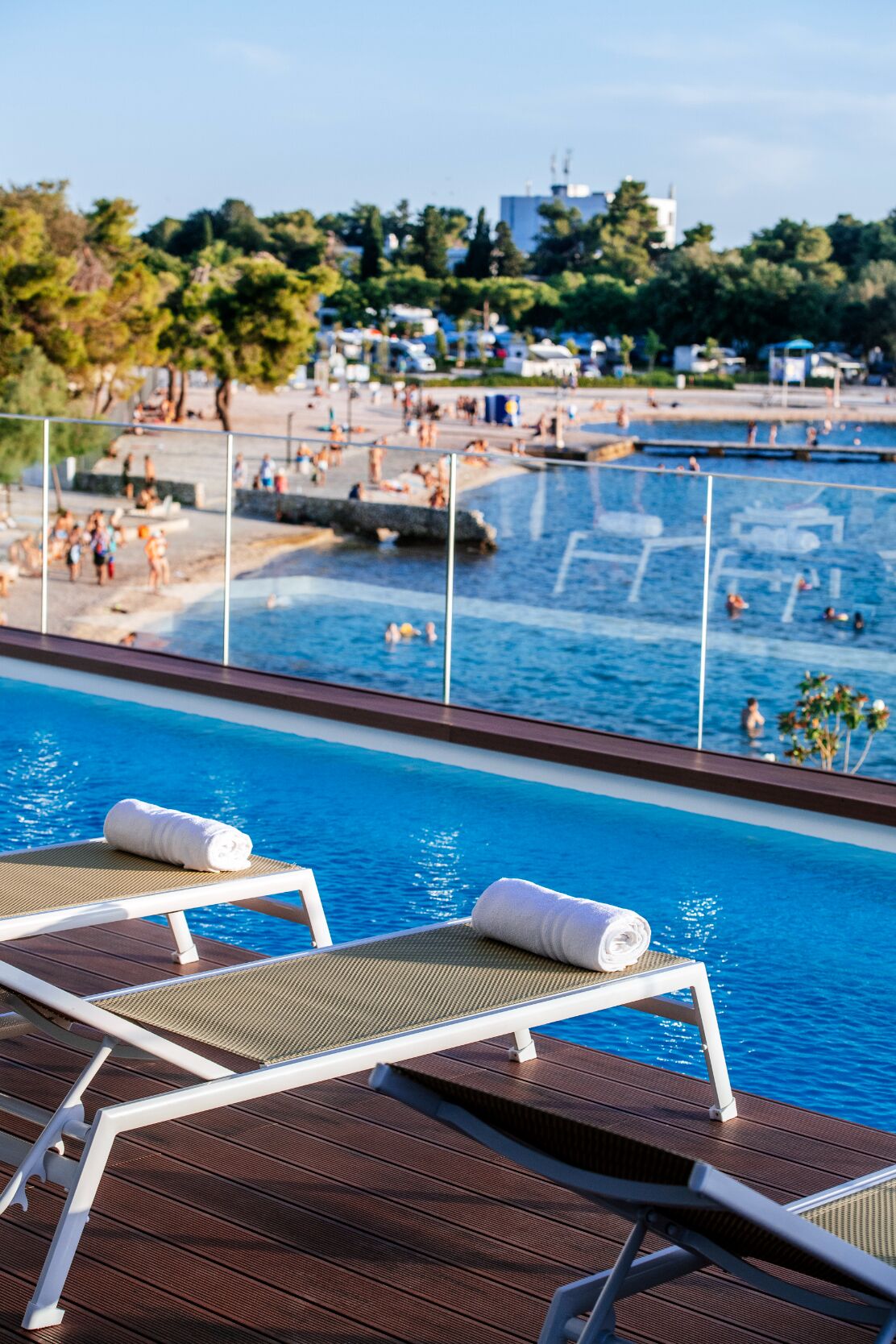 Un espace aménagé au bord de la piscine, avec des chaises longues et une vue sur une plage animée et les activités nautiques qui s’y déroulent.