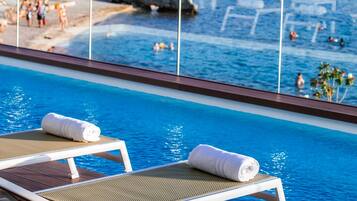Un espace aménagé au bord de la piscine, avec des chaises longues et une vue sur une plage animée et les activités nautiques qui s’y déroulent.