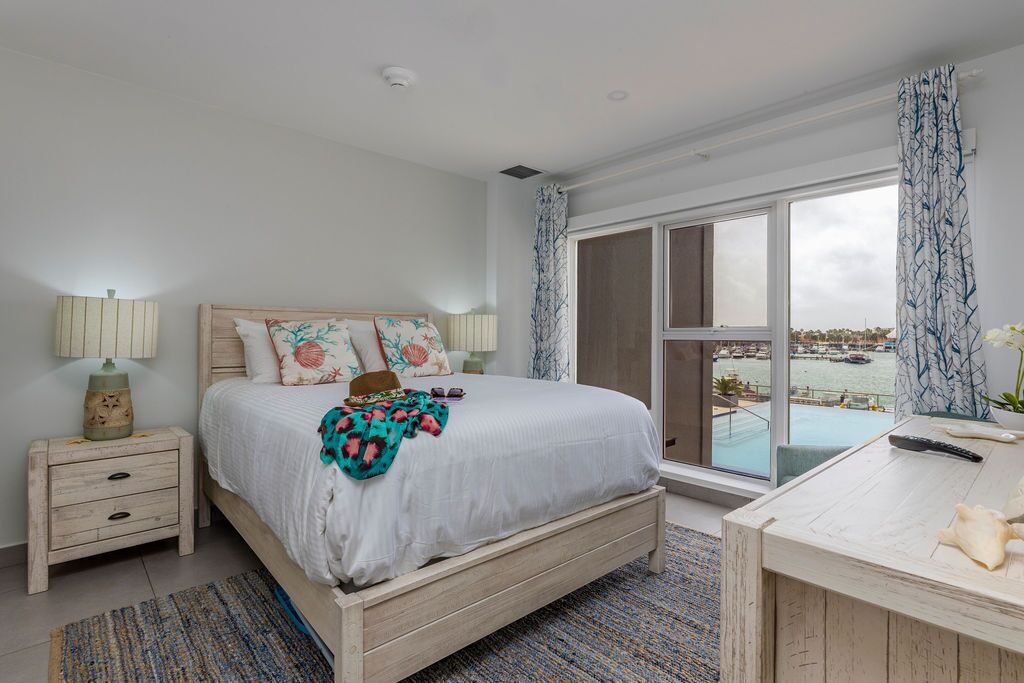 A bedroom with a large bed, a nightstand, a desk, and a view of the sea through a sliding glass door.
