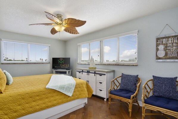 A bedroom with a bed, a desk, two chairs, a ceiling fan, and a TV.