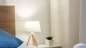 Una cama bien hecha con ropa de cama azul, una almohada blanca, una mesita de noche con lámpara, una planta en maceta y toallas enrolladas.