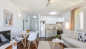 Una sala de estar moderna con televisor de pantalla plana, muebles blancos, un comedor y una cocina con gabinetes blancos y una estufa.