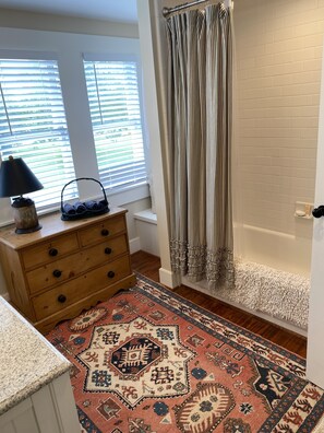 A bathroom with a shower curtain, a wooden dresser, a patterned rug, and a window with blinds. - Farm House Inn - Quiet Country Elegance (New Ulm)