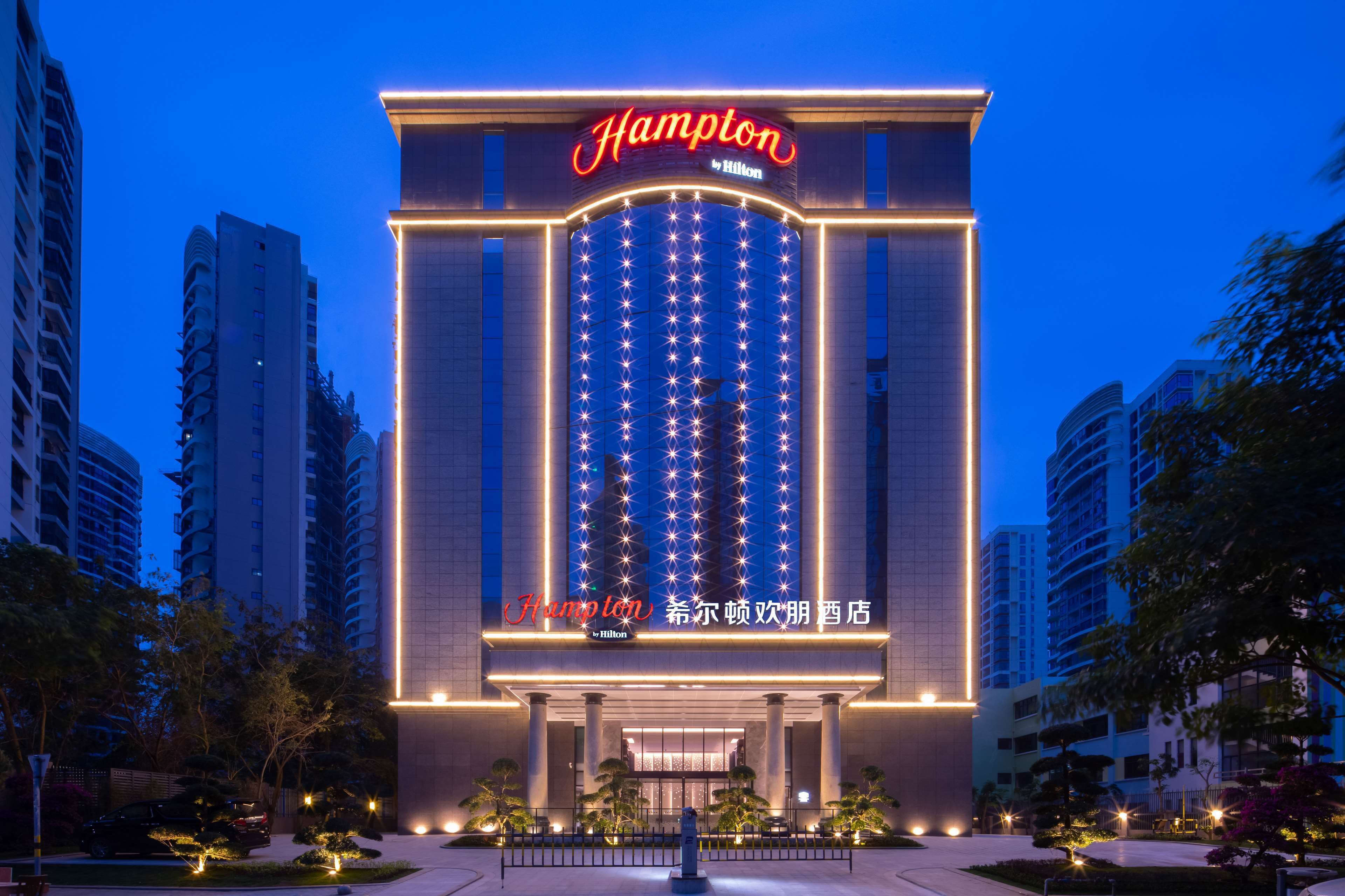 A hotel with a prominent 'Hampton by Hilton' sign, large windows, and a well-lit entrance.